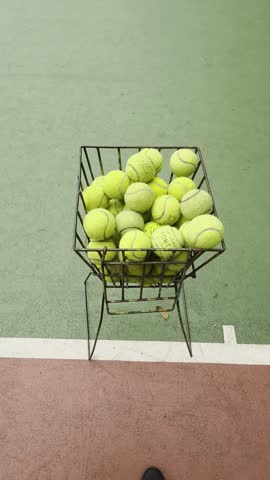 a green tennis court, featuring a wire metal basket filled with piles of yellow tennis balls in the lower right foreground.