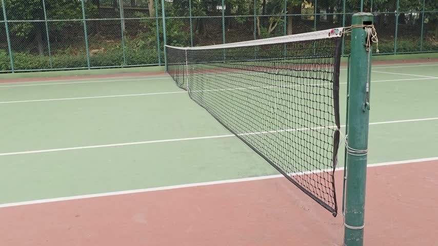 a green tennis court, featuring a wire metal basket filled with piles of yellow tennis balls in the lower right foreground.