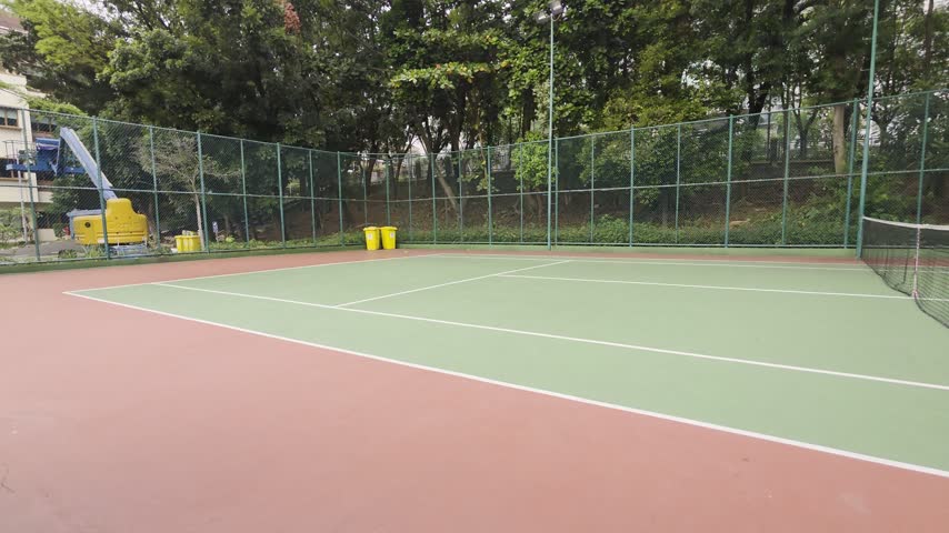 a green tennis court, featuring a wire metal basket filled with piles of yellow tennis balls in the lower right foreground.