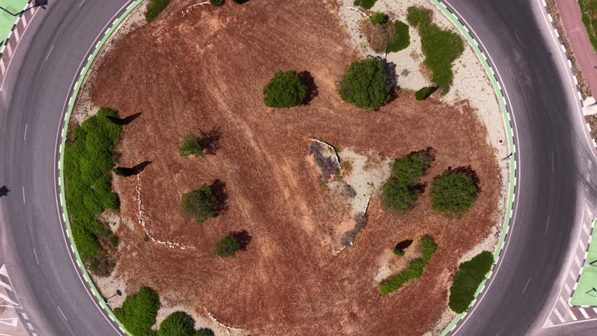 Top-down view of a circular roundabout with a landscaped center island featuring dry soil, scattered green shrubs, and cars navigating the road