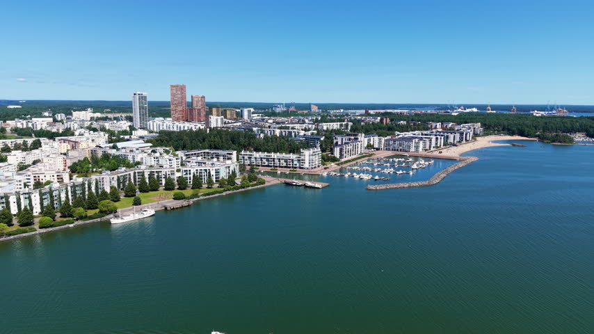 Vuosaari district in Helsinki, Finland, showing residential buildings along the Baltic Sea coastline with a marina, sandy beach, and a small boat crossing calm green waters