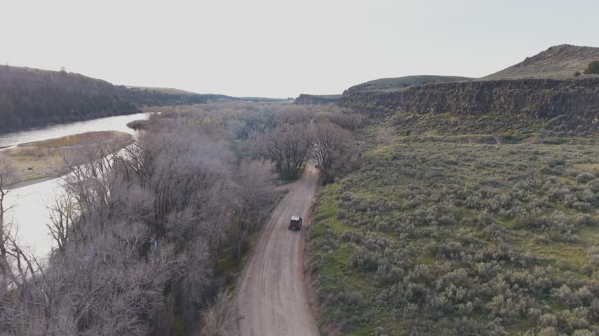 Aerial high follow of vehicles on a dirt road through bare trees and sagebrush toward a basalt canyon rim and river, Idaho