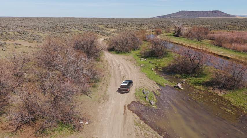 Aerial push in from a wide high desert view with a creek and butte down to a pickup truck on a dirt trail, Idaho