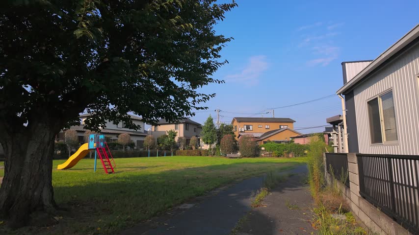 POV walking along a small path next to a neighborhood park in a Japanese residential area on a sunny morning.