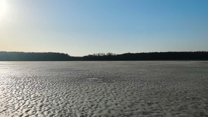 A wide view shows a frozen lake in the afternoon. The sunlight reflects off the icy surface, creating a bright scene. Nearby trees form a silhouette against the clear blue sky. No people are present. The area appears calm, and the icy landscape stretches across the foreground.