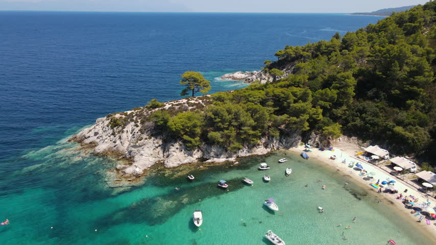 Secluded beach cove with anchored boats in crystal clear turquoise water surrounded by rocky coastline and dense Mediterranean pine forest. Smooth aerial tracking shot moving left to right along.
