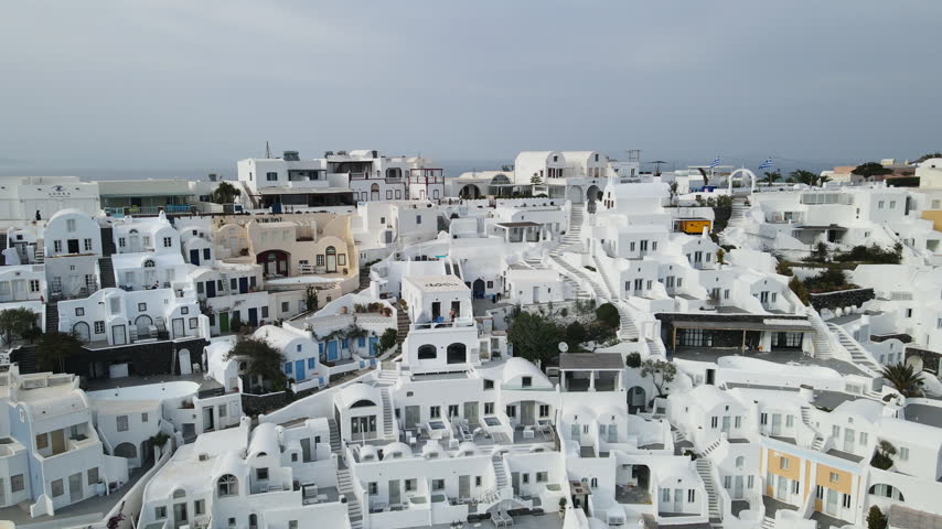 Traditional whitewashed buildings with blue accents cascade down the clifftop village of Fira in Santorini, Greece. Smooth descending aerial shot reveals terraced hotels with swimming pools.