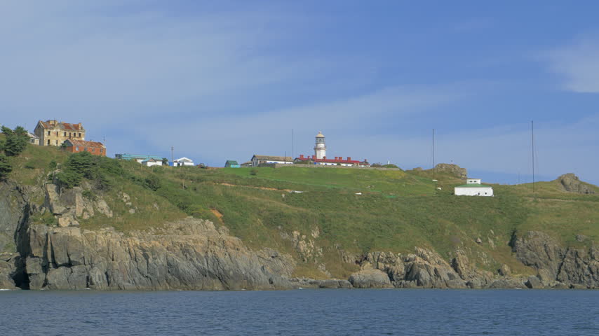 Primorye lighthouse station spreads across a grassy rocky cape with a white beacon tower red roof service buildings antenna masts and weathered houses viewed from calm coastal water in Primorye