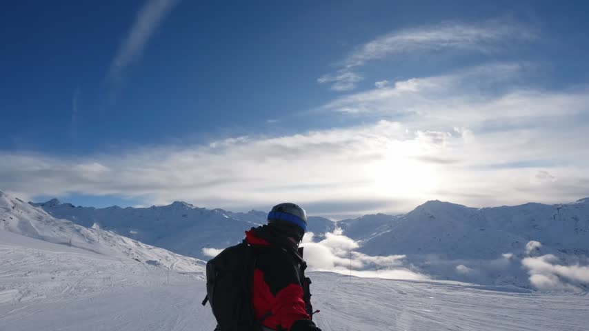Rear three-quarter view of a snowboarder with a backpack facing snowy mountains and low valley clouds in bright winter backlight.
