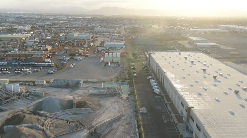 Steady aerial camera glides beside a warehouse service road with loading bays and an adjacent industrial materials yard.