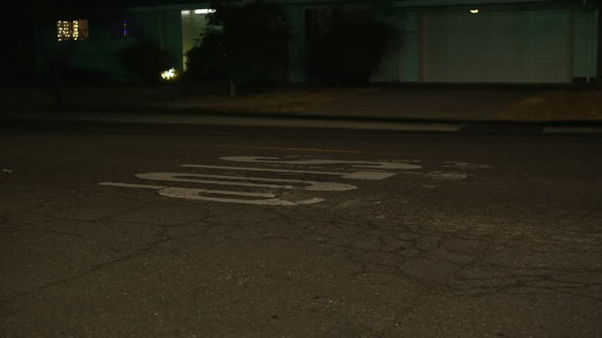 STOP road marking on a dark residential street late at night is visible on cracked asphalt before an intersection, with a dimly lit suburban house in the background