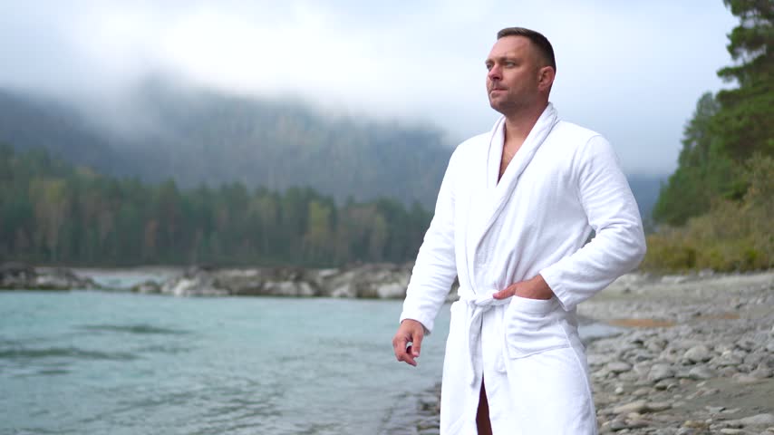 Handsome young man in a white bathrobe standing on a rocky riverbank, looking at a fast mountain river on a foggy morning.