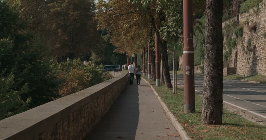 Quiet pedestrian path beside a historic stone wall in Bergamo, Italy, lined with mature trees and autumn leaves, with a road and traditional city architecture nearby.