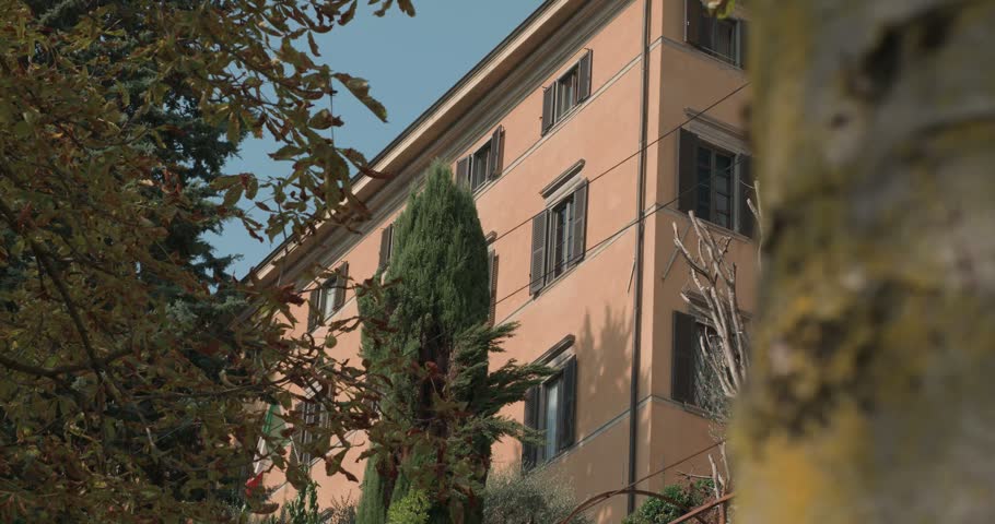 View of a traditional warm-toned building in Bergamo, Italy, partially framed by mature trees and leafy branches. The façade features multiple windows with dark shutters and a sloping roof, while surrounding vegetation creates a layered composition between the architecture and the natural environment. An architectural detail shot capturing the character of the historic urban landscape in Bergamo