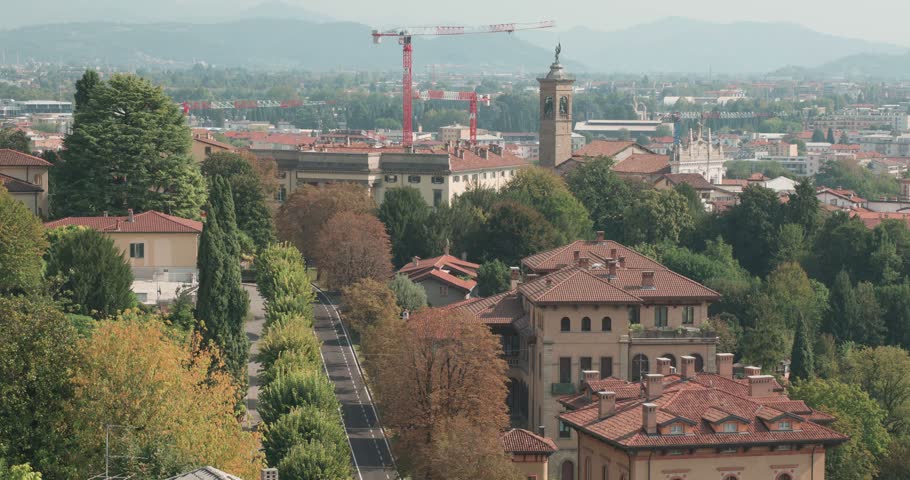 Elevated panoramic view across Bergamo, Italy, looking over residential buildings, terracotta rooftops and tree-lined streets. A prominent red construction crane and a historic tower rise above the surrounding cityscape, while distant buildings extend towards the hazy horizon. The footage captures the contrast between traditional Italian architecture, urban development and green spaces in this historic city in Lombardy.