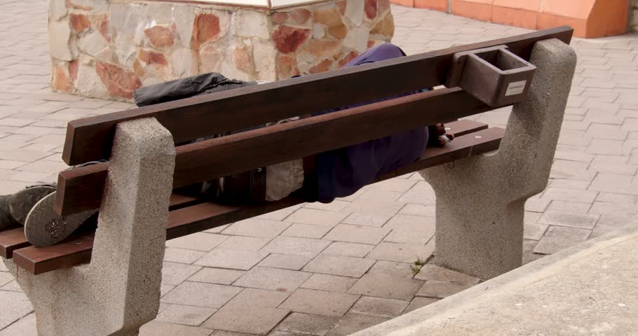 Poor homeless man or refugee sleeping on the wooden bench on the urban street in the city.