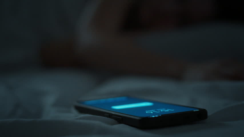 A young woman extends her hand toward an illuminated mobile phone in the dark room. The lit device displays a notification interface, representing modern technology use during nighttime hours