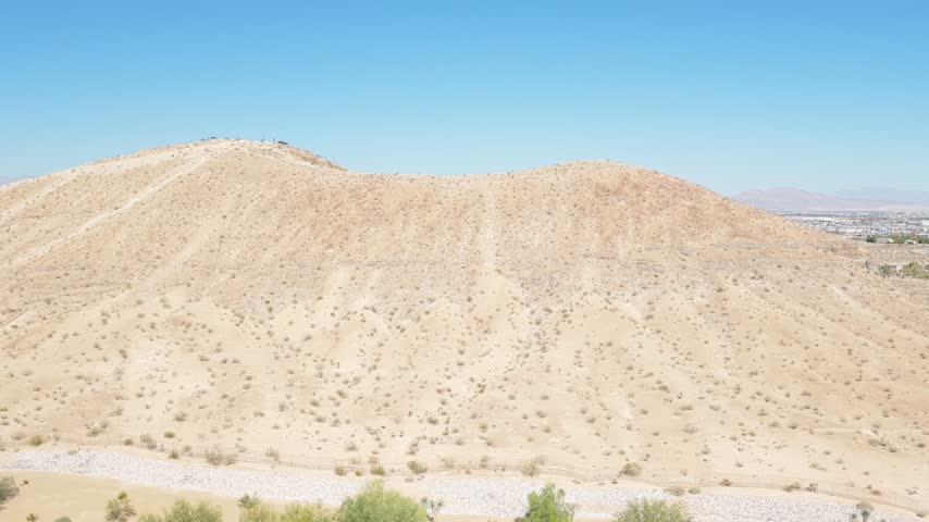 Stable drone view frames a rugged barren hill rising above green trees beneath a deep blue sky.