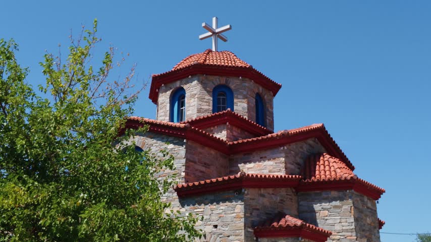 Greek Orthodox church with traditional red roof and bell tower surrounded by trees in Thassos, Greece, under a clear summer sky.