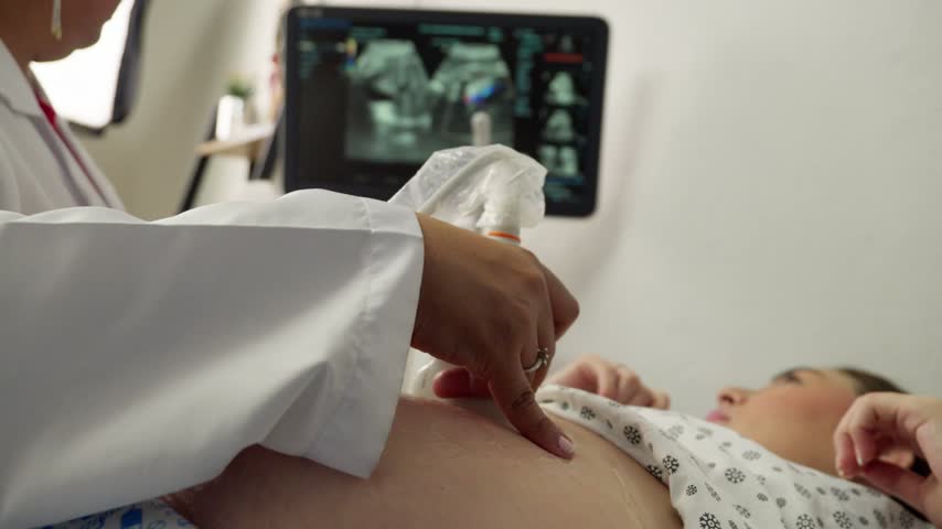 A young pregnant woman of Latina descent looks at the screen during a routine nine-month ultrasound scan. The medical professional positions the transducer on her belly to view the baby.