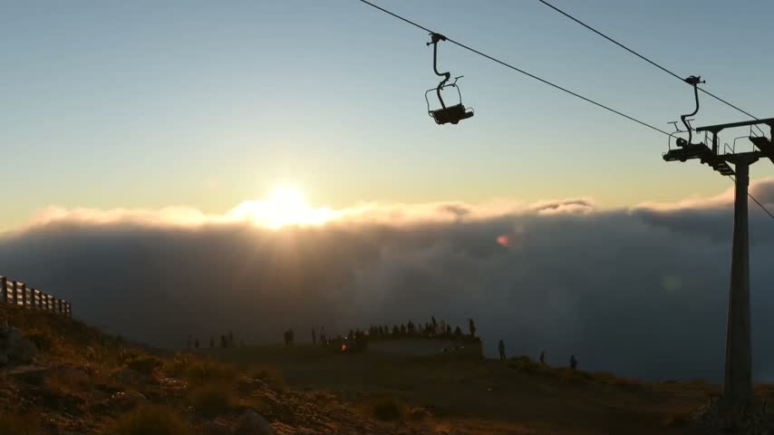 Time-lapse footage of a sunset over a mountain ridge above a sea of clouds, featuring a ski lift and silhouetted people gathering at a viewpoint.