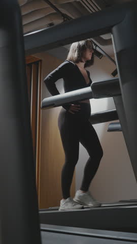 Pregnant woman walks at a steady pace on a treadmill during a prenatal cardio workout in a modern gym in vertical slow motion.