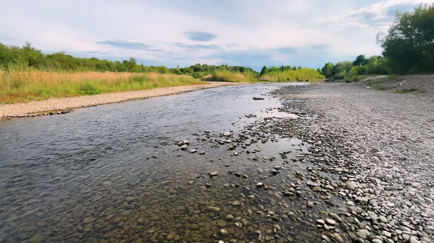 Shallow mountain river with a pebble bank flows under a cloudy sky. Crystal clear stream runs through a wild valley in summer. Picturesque view of the rocky shores and green forests. Natural water landscape in the daytime.