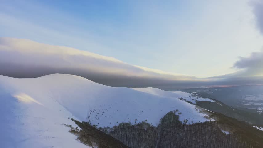 Cloud waves gently and slowly flow over the tops of snow-capped mountains covered with spruce forests at a cold sunset