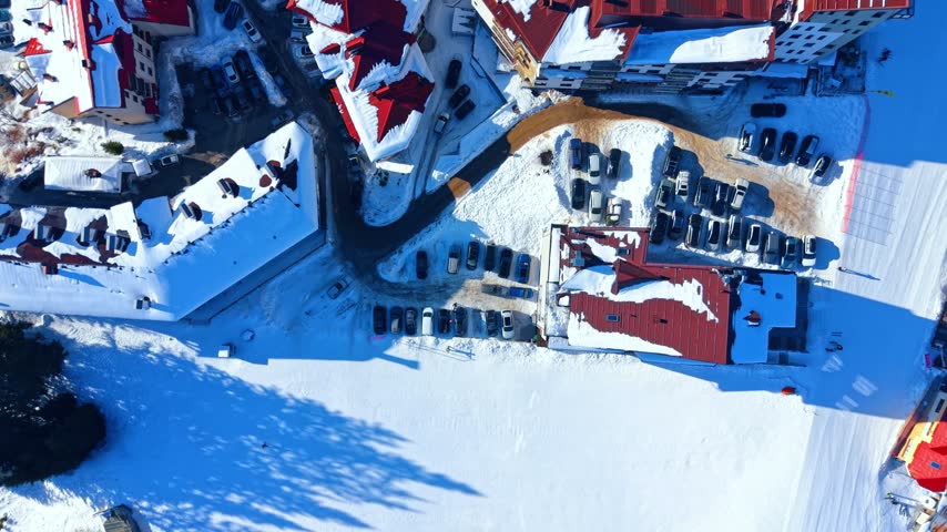 Top view of hotel parking lot and roofs in a winter resort. Aerial shot shows rows of parked cars between buildings with red roofs on snow ground.