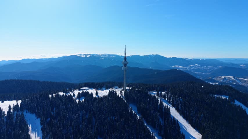 Telecommunication tower on snowy mountain top above pine woods. A tall concrete structure stands among dark green trees under blue sky.
