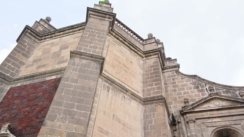 A stone Baroque church facade with a rust red cross mounted beside a gated entrance, featuring tezontle volcanic stone detail.