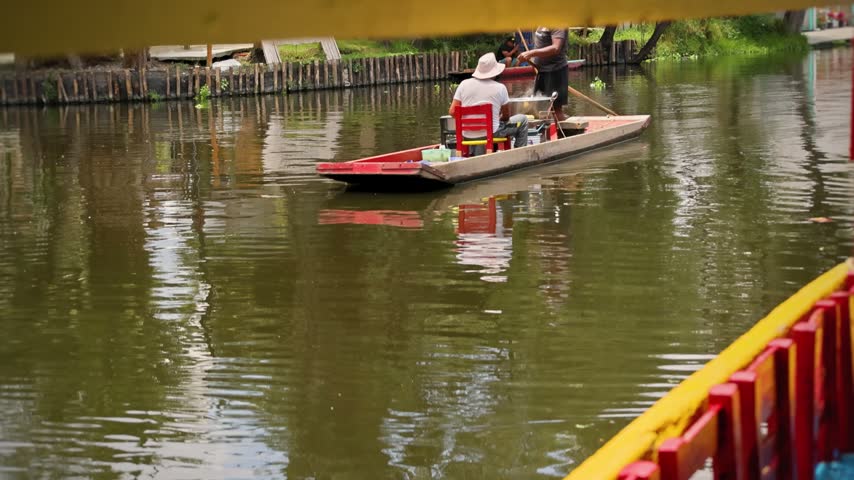 A wooden canoe with two men and a small vendor setup glides along the Xochimilco canal in Mexico City.