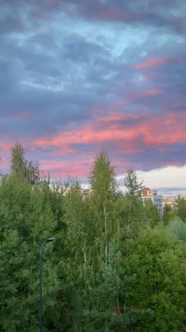 Vilnius, Lithuania - September 10 2026:  A vertical scenic photograph capturing a colorful sunset sky over a suburban park and green space. The image displays a dramatic cloudscape filled with soft pink, purple, and blue tones illuminated by the setting sun during twilight. Below the sky, lush green treetops, birch leaves, and wild vegetation occupy the foreground and middle ground, with distant residential apartment buildings and utility lines subtly visible along the horizon. The tranquil atmosphere highlights the harmony between urban residential areas and natural green zones in the evening. Ideal for stock photography related to nature landscapes, twilight skies, background textures, mobile screens, urban greenery, and environmental themes.