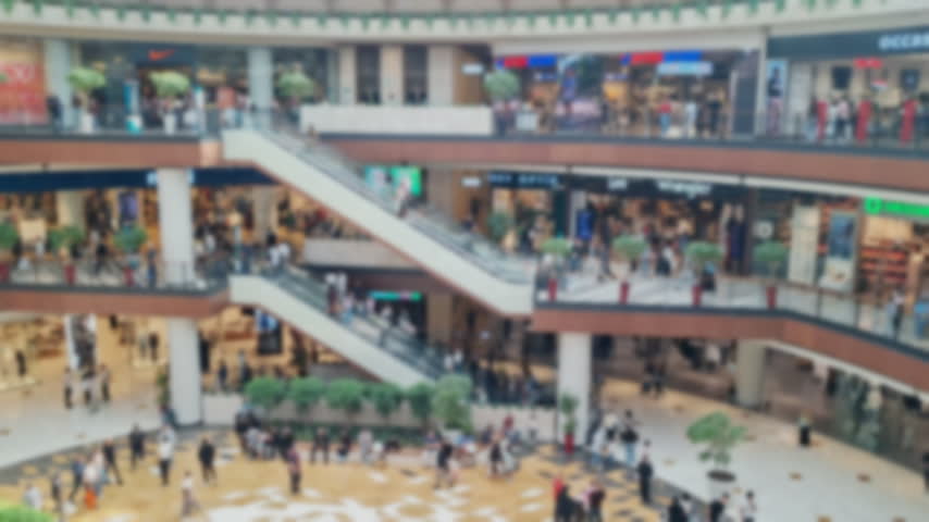 Blurred shopping mall interior showing shoppers, retail stores, escalators, and a lively commercial atmosphere with soft focus and abstract movement.