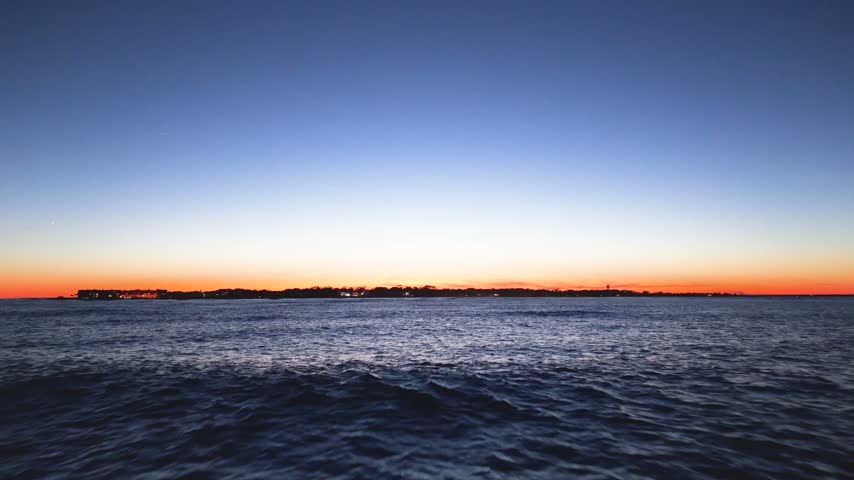 Low-altitude aerial view skimmed over ocean water toward a distant coastal island silhouette against a vibrant orange and blue sunset horizon in South Carolina.