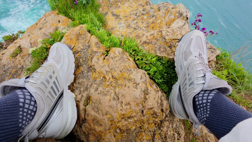 POV sitting on a rocky cliff edge above crashing turquoise surf with legs in white trousers beside purple wildflowers, tilting up to the open sea at Cabo Mayor, Santander, Cantabria, Spain