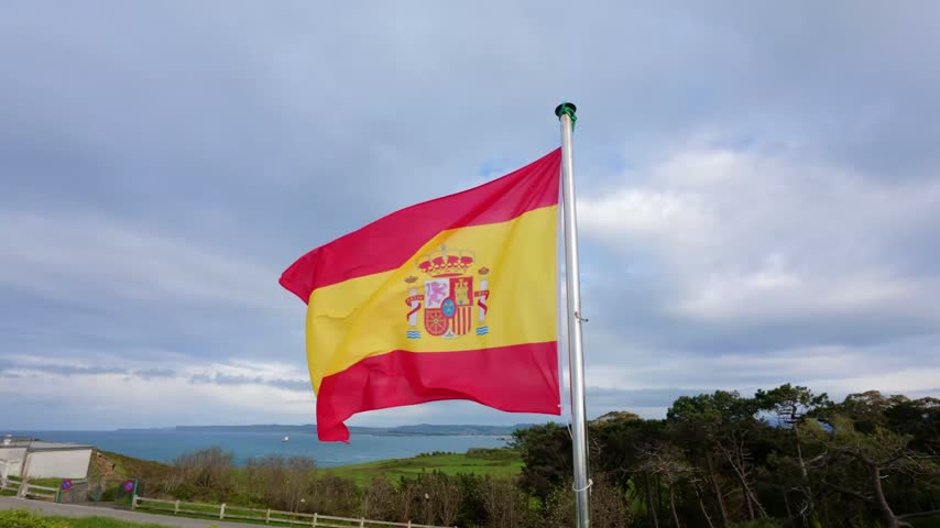Spanish national flag with coat of arms waving in slow motion on a flagpole above a green headland at Cabo Mayor with the sea and coast beyond under a grey cloudy sky, Santander, Cantabria, Spain