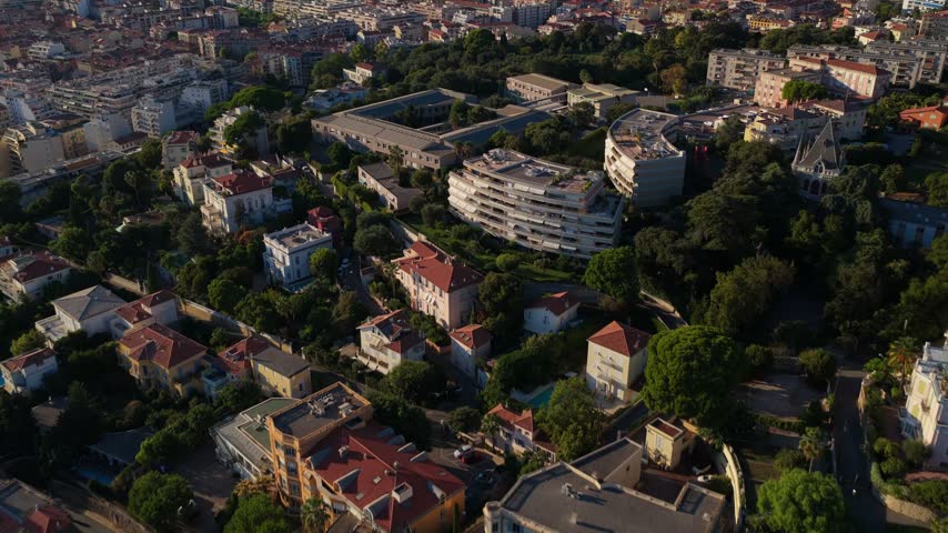 Forward flight over villas and a green park, tilting up to reveal dense city rooftops and hills in warm afternoon light, Nice, French Riviera, Cote d Azur, France