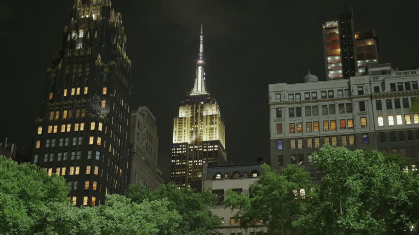 The Empire State Building glows above the green trees of Bryant Park at night between an Art Deco tower and lit office windows. Dark city sky over Midtown Manhattan in New York City, New York, USA.