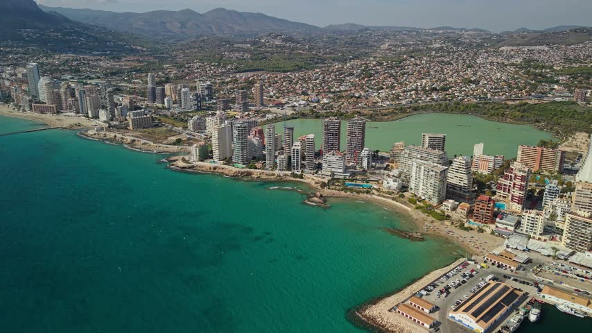 Drone flying over turquoise ocean water next to beach resort town with many tall buildings and a lake.