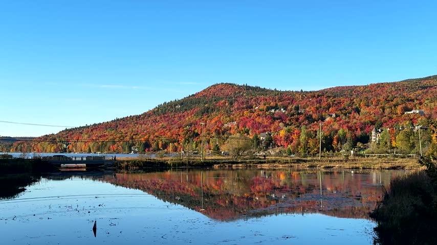Scenic view of a colorful autumn mountain reflected in calm water in Mont Tremblant village, Quebec, Canada, with red, orange and yellow forest foliage under a clear blue sky.