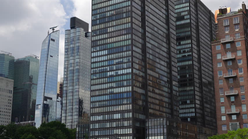 Chrysler Building spire appears between glass skyscrapers of Midtown East in New York City, New York, USA. Cloudy summer sky, dark office tower and green trees in front. Moving camera reveal.