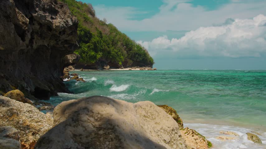 Waves crash against rocky coastline while soft sand stretches along the shoreline. Lush hills rise above crystal clear water. Bright blue sky enhances the vibrant scenery.