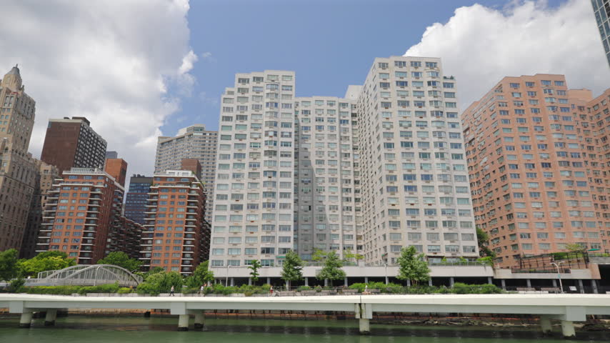 White esplanade on piers runs along the East River below apartment towers in New York City, New York, USA. Arched footbridge, green trees, walkers and summer clouds in blue sky on a sunny day.
