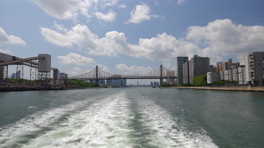 Ferry wake churns white foam on East River toward Queensboro Bridge in New York City, New York, USA. Roosevelt Island apartments on the right, industrial conveyor on the left, sunny sky with clouds.