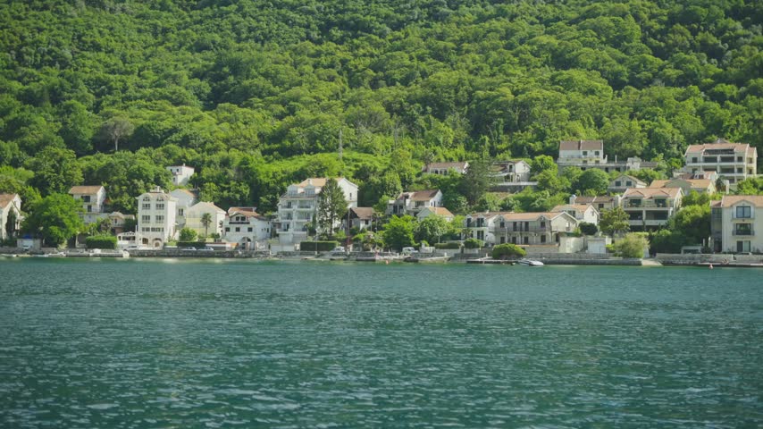 coastal village houses built into lush green mountain slopes overlooking calm blue Adriatic sea waters in Montenegro during bright sunny day showing Mediterranean residential architecture