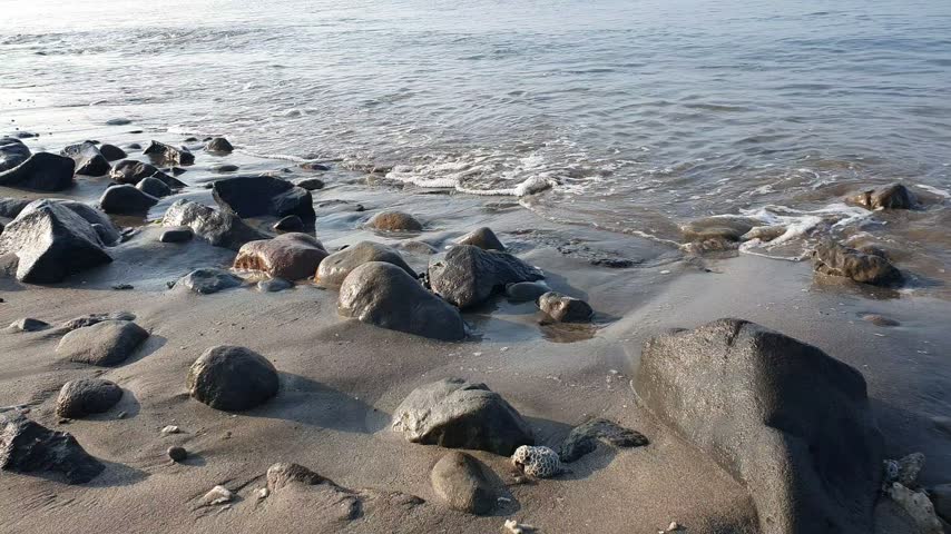 Close-up shot of gentle ocean waves softly washing over a sandy shoreline filled with scattered rocks and stones.