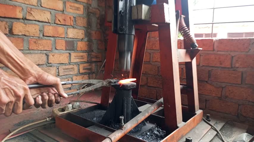 SOUTH SUMATRA, INDONESIA - OCTOBER 2026: Blacksmith using a mechanical power hammer machine to forge and shape a hot glowing piece of iron in a traditional workshop.