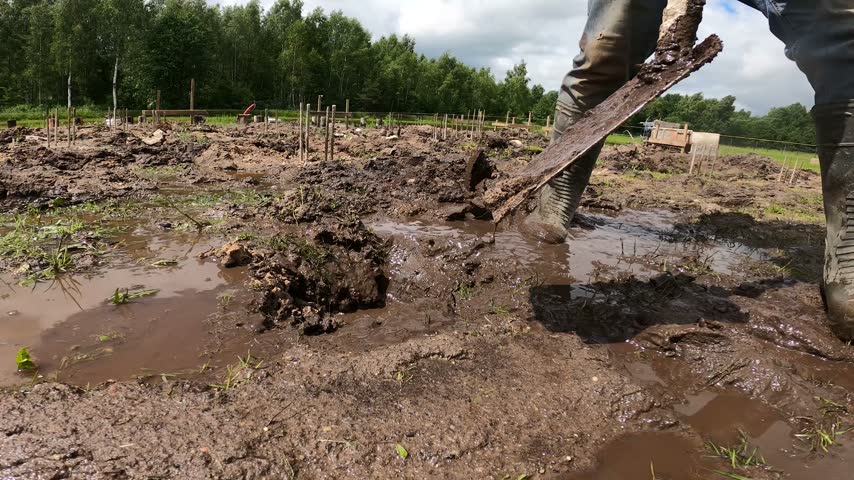 Low-angle view of an unrecognizable worker repeatedly pressing a shovel into wet mud and lifting soil during foundation groundwork.