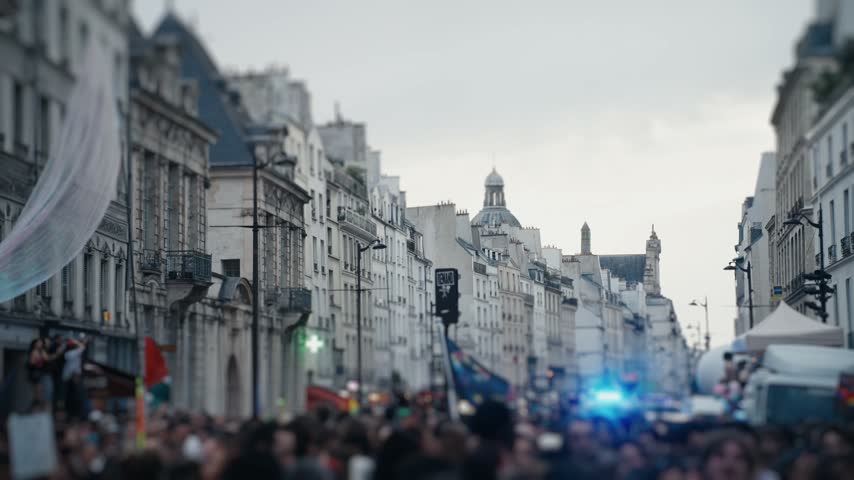 Large crowd of protesters marching on a european city street with police lights flashing in the background